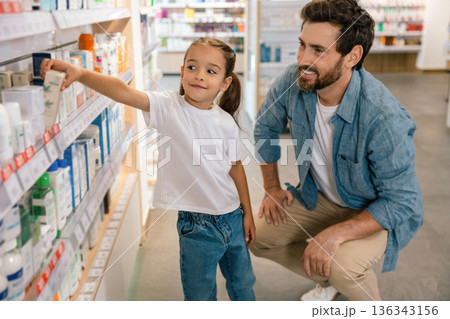 Father squatting next to daughter reaching for product on shelf in pharmacy 136343156
