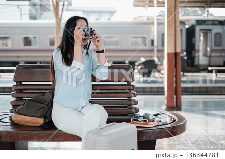 Woman sitting on bench at train station, taking photos with vintage camera. Travel and adventure concept. 136344781