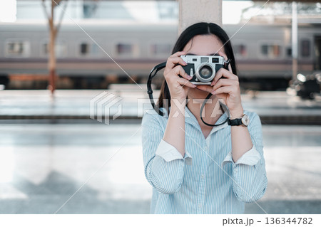 A woman in a blue shirt holds a vintage camera at a train station, ready to capture memories. A woman in a blue shirt holds a vintage camera at a train station, ready to capture memories. 136344782