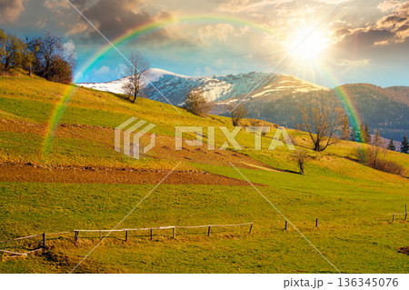 remote mountainous rural landscape in springtime at sunset. arable and trees on steep slope in green grass in evening light. distant snow capped peak. april weather under rainbow and clouds on the sky remote mountainous rural landscape in springtime at sunset. arable and trees on steep slope in green grass in evening light. distant snow capped peak. april weather under rainbow and clouds on the sky 136345076