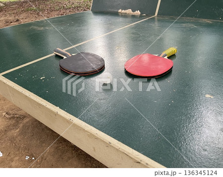 Two table tennis paddles and a ball are resting on a green table in a park during the afternoon 136345124