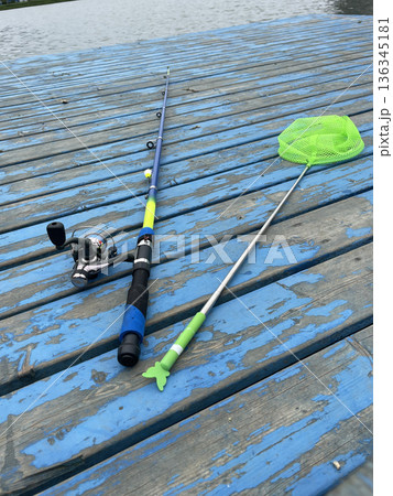 Fishing equipment on wooden dock by water at sunset with fishing rod, reel, and net ready for use 136345181