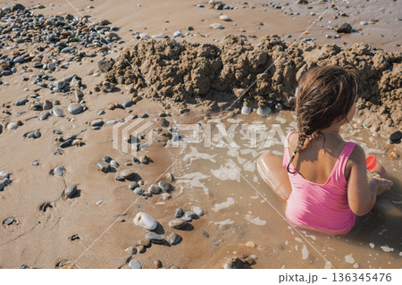 Little girl in a pink swimsuit playing actively with a red toy on the wet sand, building a moat and fort on a sunny pebble beach during a summer vacation 136345476