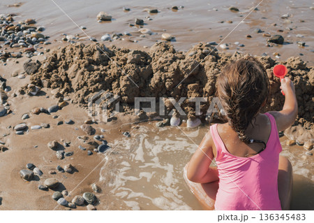 Little girl in a pink swimsuit playing actively with a red toy on the wet sand, building a moat and fort on a sunny pebble beach during a summer vacation 136345483