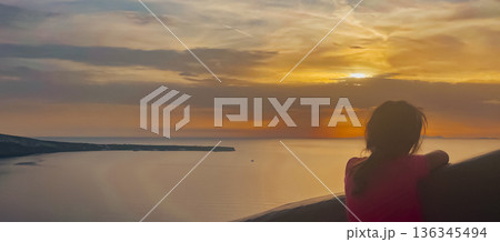 Silhouetted figure of a child against the colorful golden-hour sky in the picturesque cliffside village of Santorini. 136345494