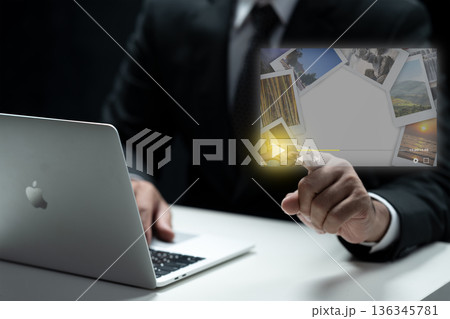 A businessman in a suit uses a laptop while interacting with a holographic video player interface featuring photo thumbnails. Concept for digital marketing, video streaming, and future technology 136345781