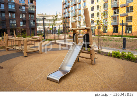 Modern outdoor playground with a metal slide and wooden bridge structure on a safe rubberized surface for child play. Residential area with new architecture. Modern outdoor playground with a metal slide and wooden bridge structure on a safe rubberized surface for child play. Residential area with new architecture. 136345818