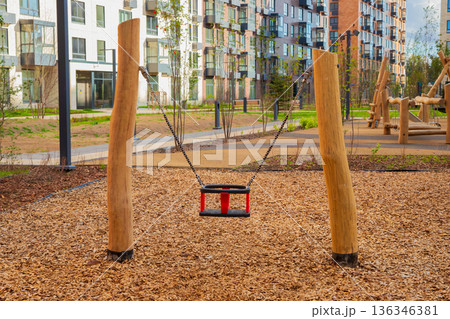 Empty baby swing on a modern wooden playground with a soft wood chip surface and apartment buildings in the background for children recreation. Empty baby swing on a modern wooden playground with a soft wood chip surface and apartment buildings in the background for children recreation. 136346381
