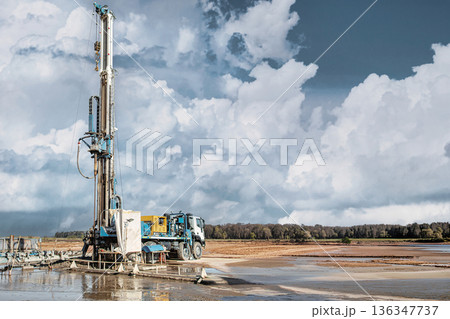 A drilling rig is set up to extract oil or gas in an outdoor area. The rig stands tall against a cloudy sky while the landscape shows water and dry ground A drilling rig is set up to extract oil or gas in an outdoor area. The rig stands tall against a cloudy sky while the landscape shows water and dry ground 136347737