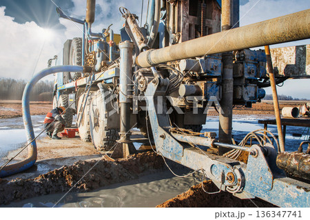 Workers operate a drilling rig on a site to extract oil and gas. The equipment is set up on the ground with various tools and machinery in use Workers operate a drilling rig on a site to extract oil and gas. The equipment is set up on the ground with various tools and machinery in use 136347741