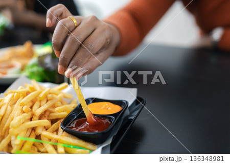 Close up of female hand holding a golden french fry and dipping it into tomato ketchup. Fast food snack on tray with cheese dip. Casual lifestyle eating in a restaurant or cafe background. 136348981