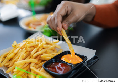 Close up of female hand dipping a golden french fry into creamy cheese sauce. Fast food snack with ketchup on tray. Casual lifestyle eating in a restaurant. Focus on delicious melted cheese dip. 136348983