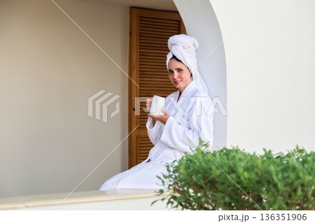 A woman in a robe and towel drinks a hot drink on a terrace after a shower A woman in a robe and towel drinks a hot drink on a terrace after a shower 136351906
