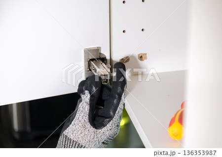 Protective gloved hand carefully installing a metal cabinet hinge into a white cupboard, representing diligent DIY project completion and home renovation work 136353987
