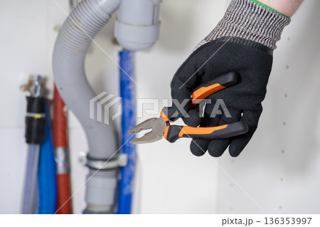 Plumber's gloved hand holding adjustable pliers near complex water pipes, ready for skilled repair and urgent maintenance work Plumber's gloved hand holding adjustable pliers near complex water pipes, ready for skilled repair and urgent maintenance work 136353997