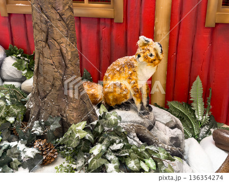 Artificial fox sitting on snowy rocks next to a tree trunk, part of a rustic winter forest display, evoking holiday and festive nature themes 136354274