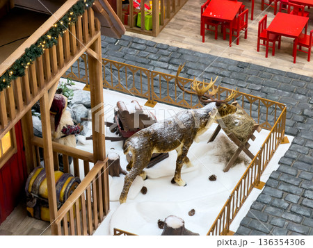 Winter holiday display with a snowy reindeer statue, sleigh, and decorated hut, evoking festive cheer in a shopping center for seasonal celebration 136354306