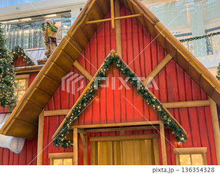 Festive red wooden house decoration adorned with garlands, string lights, and a charming figure creating a whimsical holiday spirit within a shopping mall display 136354328
