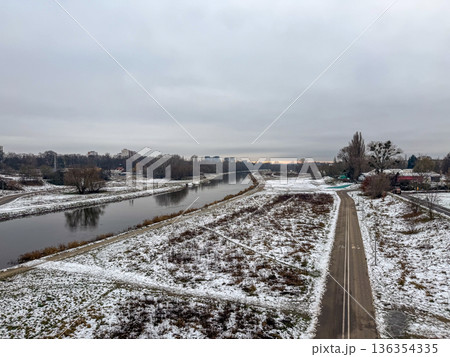 Overcast winter landscape featuring a tranquil calm river, snow-dusted banks, and a winding pathway leading toward a distant city under a grey sky, highlighting urban nature Overcast winter landscape featuring a tranquil calm river, snow-dusted banks, and a winding pathway leading toward a distant city under a grey sky, highlighting urban nature 136354335
