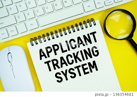 Notepad with the text "Applicant Tracking System" and white keyboard, wireless mouse, magnifying glass on yellow background 136354681