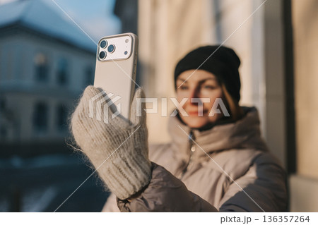Woman takes selfie outside in winter with a mobile phone while wearing a warm jacket and hat 136357264