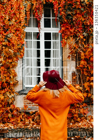 Woman in an orange sweater and a hat near a wall with wild grapes in autumn 136358421