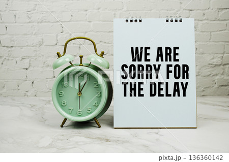 We Are Sorry For The Delay with green vintage alarm clock against a white brick wall background. 136360142