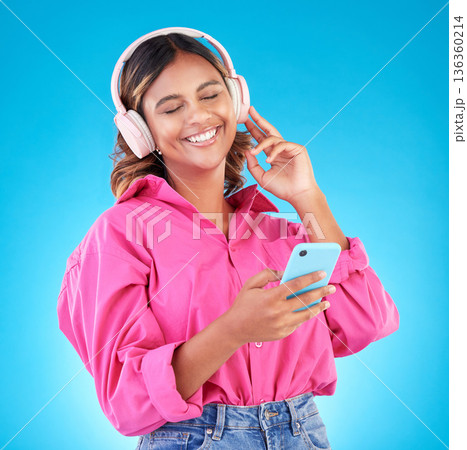 Phone, music and headphones with a woman in studio on a blue background, listening to the radio. Mobile, smile and a happy young female person streaming audio to relax for wellness or freedom Phone, music and headphones with a woman in studio on a blue background, listening to the radio. Mobile, smile and a happy young female person streaming audio to relax for wellness or freedom 136360214