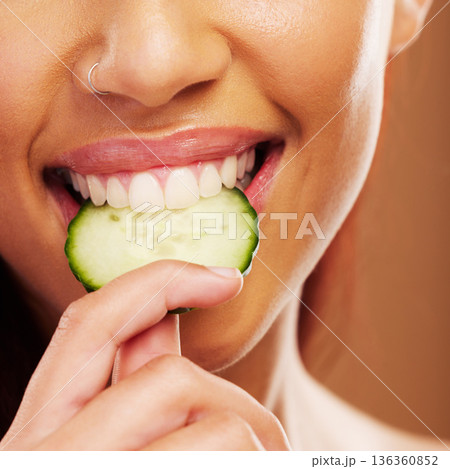 Woman, mouth or eating a cucumber for skincare nutrition or healthy diet against a brown studio background. Smile, bite or closeup of girl vegetable for organic facial treatment or natural cosmetics 136360852