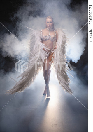 Powerful woman in white angel costume with large feather wings walking through dramatic smoke. 136362171