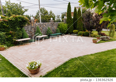 Paved patio plaza with benches in landscaped garden, stone wall, lawn and evergreen trees 136362926