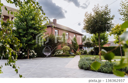 Sunlit brick house courtyard with hanging chair and manicured garden landscape 136362937