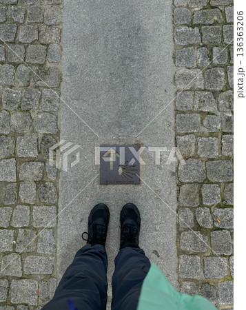 Pilgrim standing on Camino de Santiago waymark with yellow arrow and shell symbol on stone pavement. Pilgrimage, hiking journey, faith, and long-distance walking route in Europe Pilgrim standing on Camino de Santiago waymark with yellow arrow and shell symbol on stone pavement. Pilgrimage, hiking journey, faith, and long-distance walking route in Europe 136363206