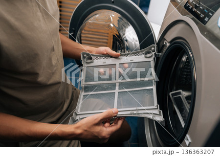 Cropped shot of man inspecting and removing lint filter from front-loading washing machine or tumble dryer, ensuring proper cleaning and essential appliance maintenance for optimal performance. 136363207