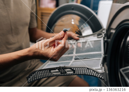 Closeup of male hand removing accumulated lint and fibers from tumble dryer filter, highlighting importance of regular cleaning for appliance maintenance and efficient operation. 136363216
