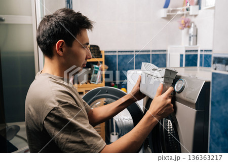 Man holding grey lint filter, removing from silver tumble dryer in laundry room, performing essential appliance cleaning and maintenance to ensure proper function and efficiency. 136363217
