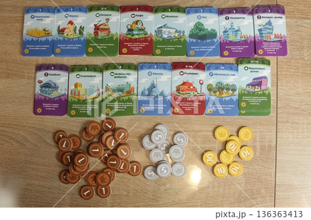 Mozyr, Belarus, April 10 2024: preparation for the board game Machi Koro. Laid out upgrade cards and coins on the table. Upper angle. Home Entertainment 136363413