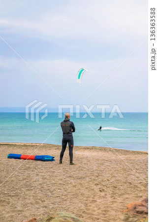Kitesurfing training on sandy beach with a man in a wetsuit watching a kitesurfing rider gliding across the sea. Concept of kitesurfing lifestyle, outdoor water sport, travel leisure, coastal activity 136363588