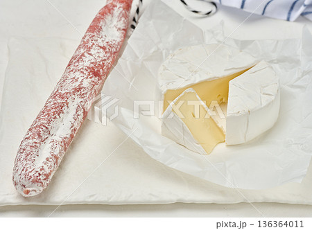 A round head of soft Camembert cheese with white mold, one triangular slice cut off, the cheese lies on crumpled white wrapping paper 136364011