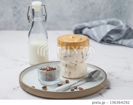 Dalgona coffee. Whipped instant iced coffee in a glass on a plate with coffee beans, bottle and spoon Dalgona coffee. Whipped instant iced coffee in a glass on a plate with coffee beans, bottle and spoon 136364292