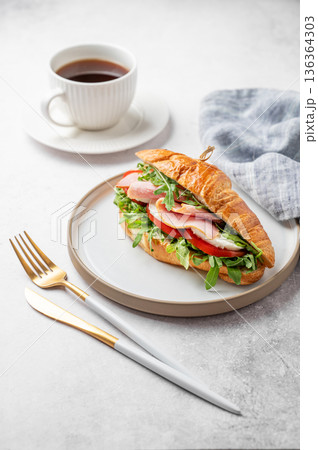 Croissant sandwich with ham, cheese, tomato, arugula on a plate and cup of coffee 136364303