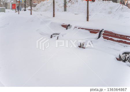 Park benches buried in snow, urban walkway after storm, snow covers seat, ground, no people. Winter city, snow accumulation, travel disruption, seasonal city services urban accessibility planning 136364867