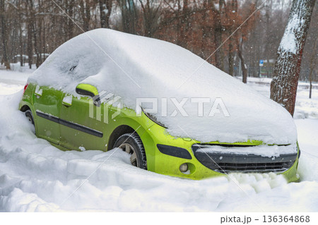 Green car parked on city street buried in snow after blizzard, windshield hidden by snow, parking blocked. Concept of snow disruption, winter transport, road safety, insurance claim 136364868