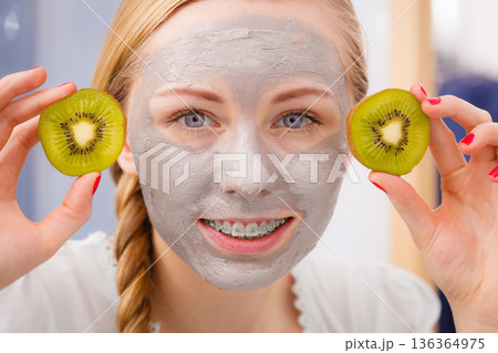 Happy young woman having face mask holding kiwi Happy young woman having face mask holding kiwi 136364975