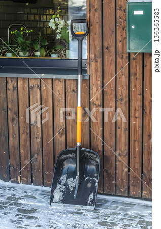 A snow shovel rests against a modern wooden facade during a light snowfall, ready for the task of clearing snow and ice from the entrance.. High quality photo A snow shovel rests against a modern wooden facade during a light snowfall, ready for the task of clearing snow and ice from the entrance.. High quality photo 136365583