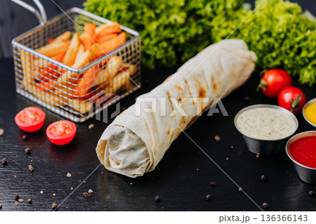 Whole shawarma wrap with fries and dipping sauces on black table 136366143