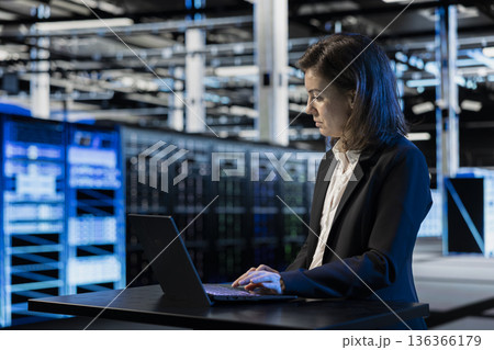Inspecting infrastructure, laptop and female programmer in data center updating network systems. IT professional using notebook to ensure stable server hub operations, overseeing rigs 136366179
