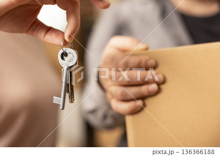 Hands of white couple presenting house keys and carrying cardboard box during relocation. Close up captures new homeowners and new beginnings in modern apartment on moving day. 136366188
