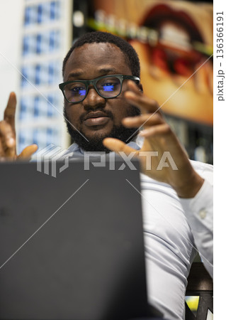 Black man participating in online video call while sitting on bench, confident street style and relaxed posture fitting naturally into the modern downtown area with vibrant active city life. 136366191