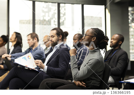 Black young female listening to presentation speech with diverse people in audience, business conference event with speaker. Chairs fill auditorium as participants attend professional summit. Black young female listening to presentation speech with diverse people in audience, business conference event with speaker. Chairs fill auditorium as participants attend professional summit. 136366195